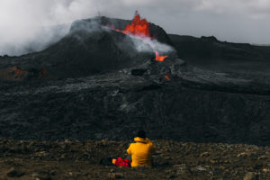 Young man backpacker in yellow jacket exploring the dramatic landscape, sitting near the big erupting volcano with melting lava
